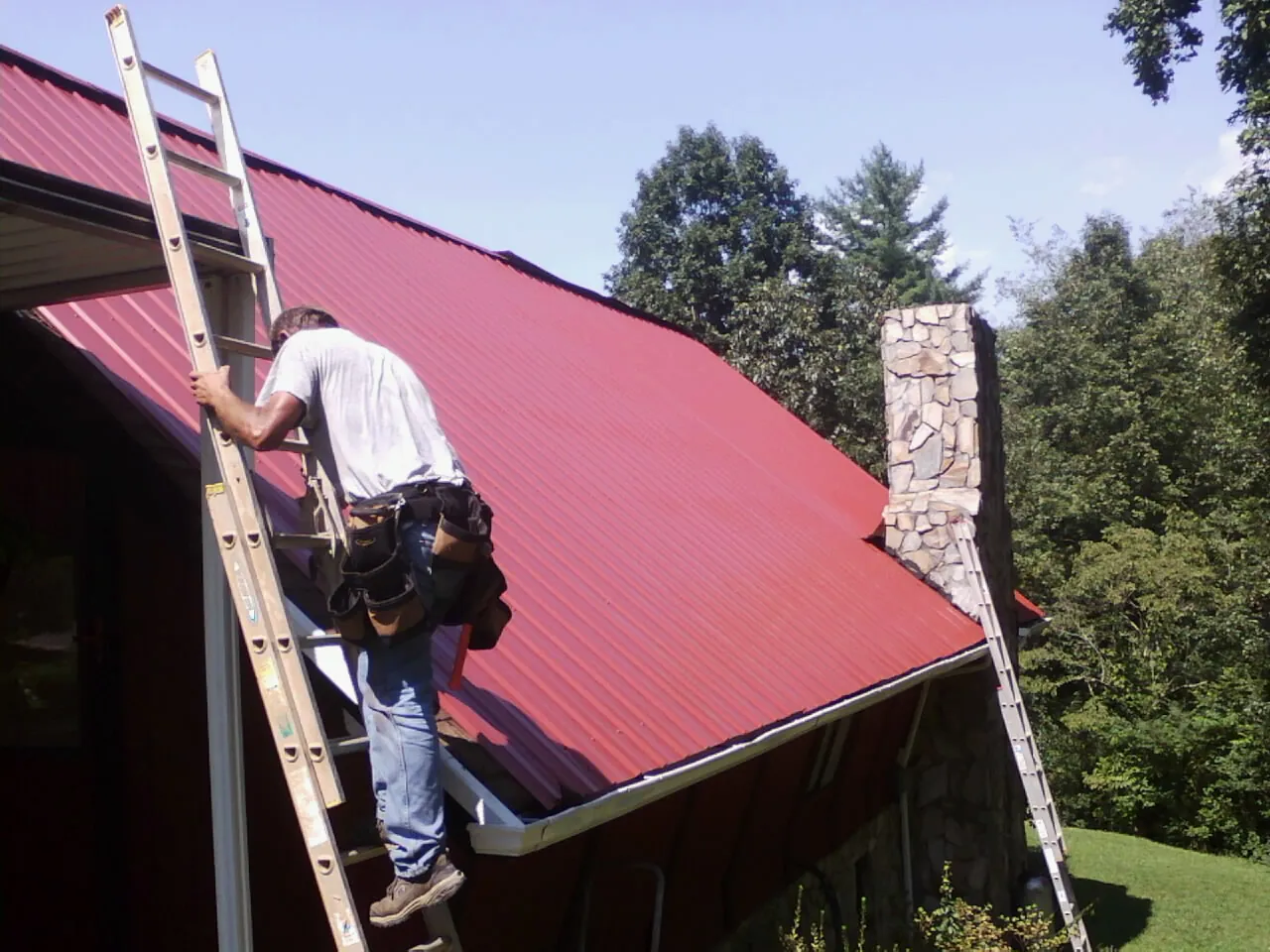 Roofer on a ladder beside a red metal roof and stone chimney — Soffit & Fascia Repair by Roofing Brothers in Pewaukee